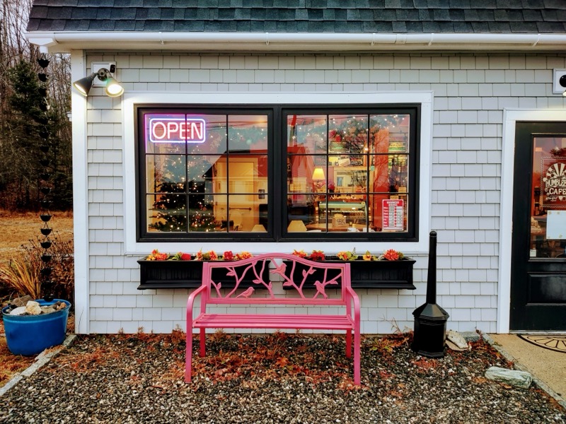 The front of the cafe: gray shingles, a lit OPEN sign in the window, flower boxes and a pink bench