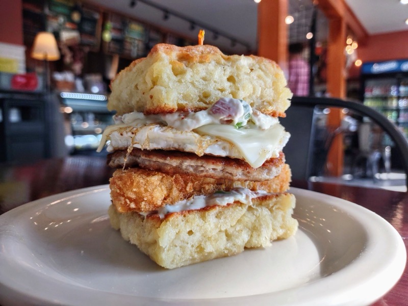 A tall breakfast sandwich on a homemade biscuit with sausage, a fried egg, cheese, a hash brown and cream cheese